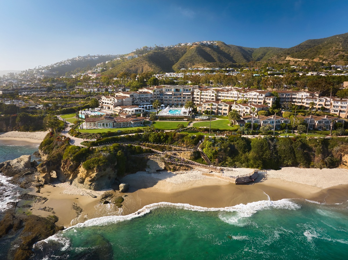 Aerial view of a large luxury resort complex perched on sandy bluffs above a turquoise Pacific cove, with a central pool terrace, manicured lawns, and multi-story guest buildings extending across the property. A wooden staircase descends the cliffside to a private crescent beach where gentle waves meet the shore. Green hillsides dotted with residential development rise behind the resort beneath a clear blue sky.