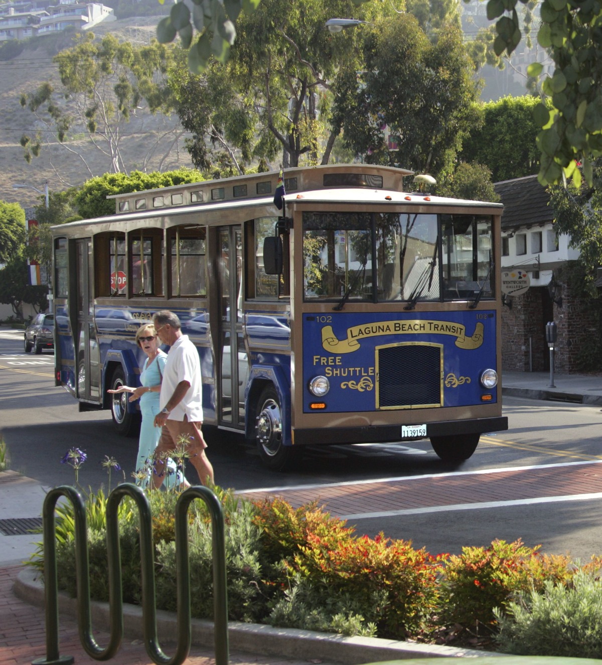 A blue Laguna Beach Transit trolley is stopped on a street lined with trees and buildings. Two pedestrians walk across the crosswalk in front of the vehicle, with greenery and a bike rack visible in the foreground.