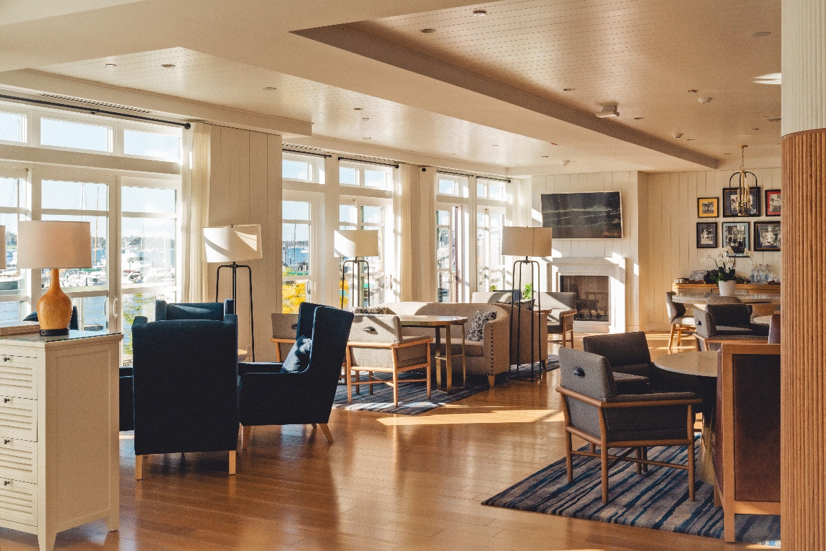 A bright, open hotel lobby with warm wood floors features an arrangement of navy and gray upholstered armchairs, floor lamps, and seating areas centered around a white fireplace. Large windows along the back wall flood the room with natural light and offer views of a marina, while framed artwork and a fluted column accent the coastal-style interior.
