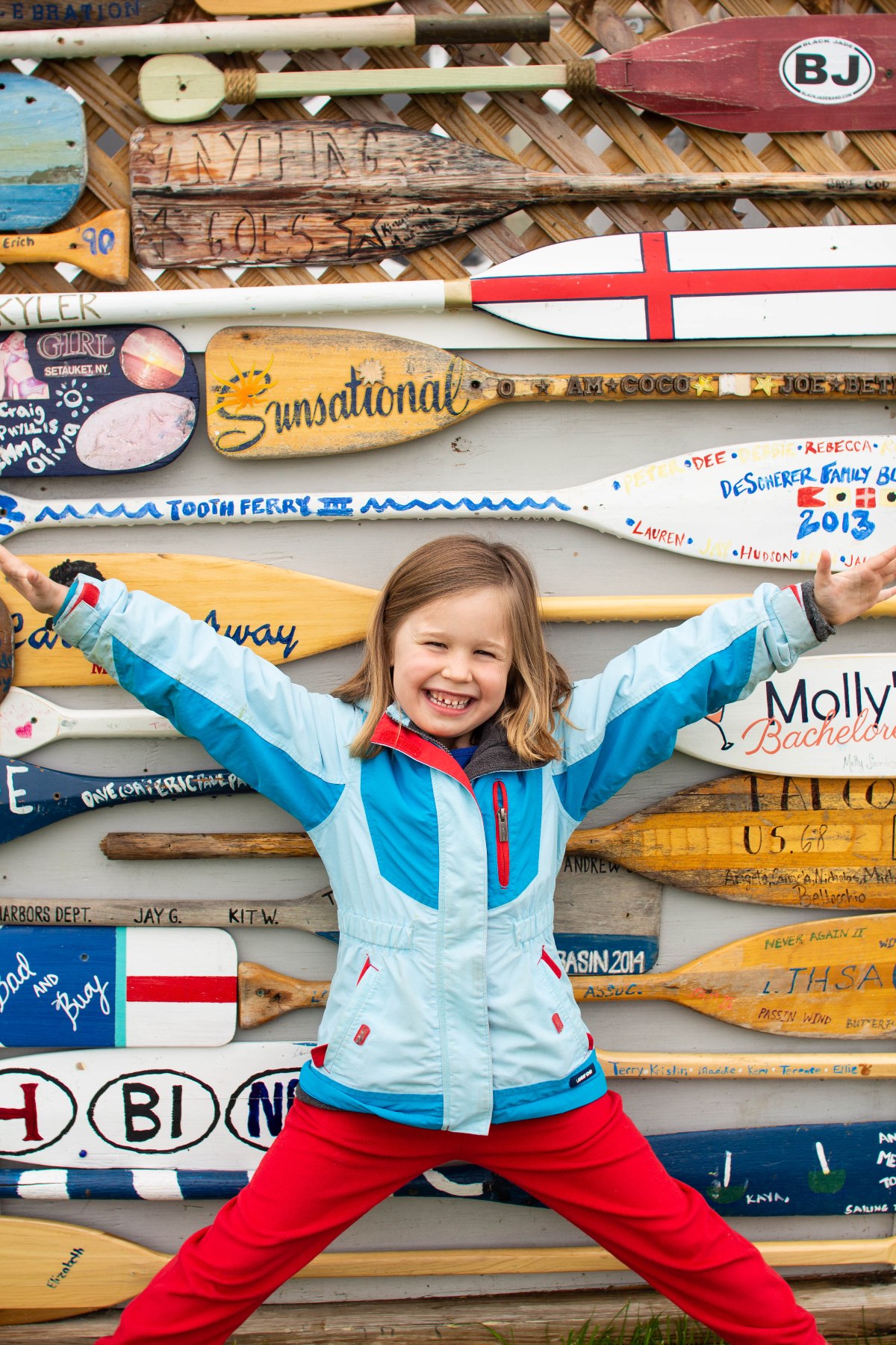 A young girl in a light blue and red jacket and red pants stands with arms and legs spread wide, smiling broadly, in front of a wall densely covered with painted and inscribed wooden oars and paddles. The oars display a variety of names, dates, flags, and colorful designs.
