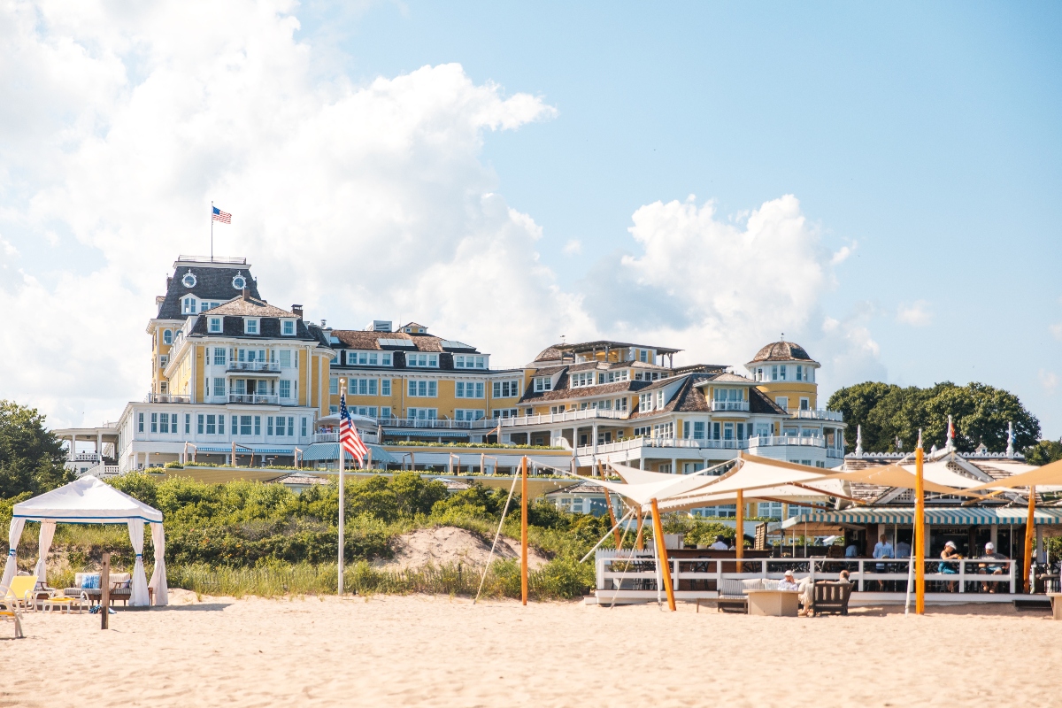 A large yellow and white multi-story Victorian-style hotel with an American flag flying from its central tower sits atop a sand dune overlooking a wide sandy beach. The beach foreground features white canvas shade structures, a gazebo, and seating areas where guests are gathered.