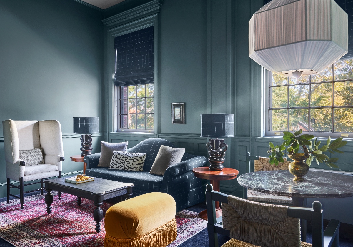 A sitting room with deep teal-painted walls and paneling features a dark plaid sofa with mixed throw pillows, a cream wingback chair, a wood coffee table, and a gold velvet fringed ottoman arranged on a red patterned rug. A marble-topped dining table, a large geometric pendant light, tall windows overlooking trees, and dark ceramic table lamps complete the richly layered space.
