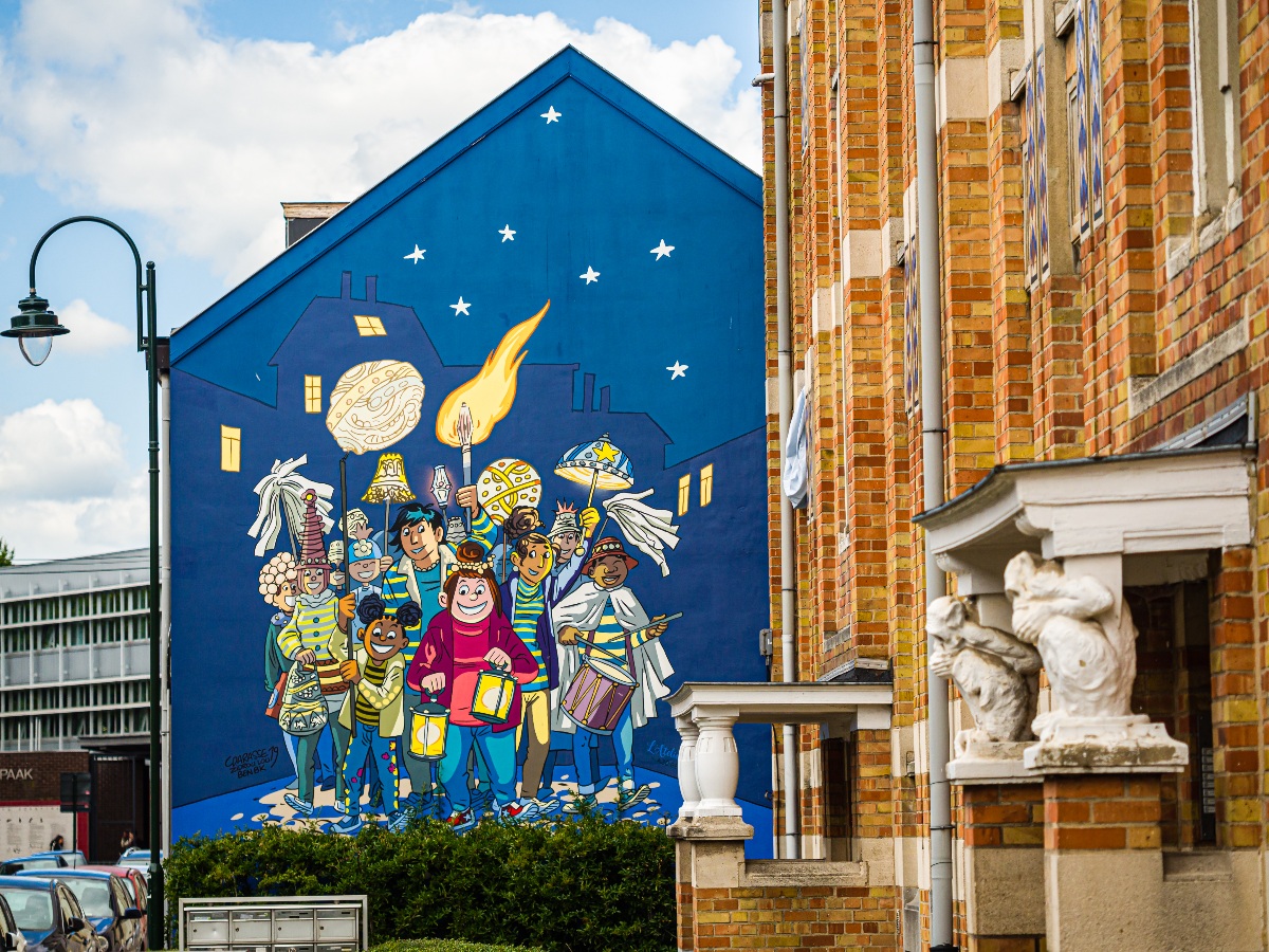 A large comic-style mural covers the blue gable end of a building, depicting a colorful group of children in festive costumes carrying lanterns, torches, and drums beneath a starry night sky with a comet overhead. The mural is flanked by a red-brick building with white classical architectural details, including a stone sculptural figure mounted at the corner.