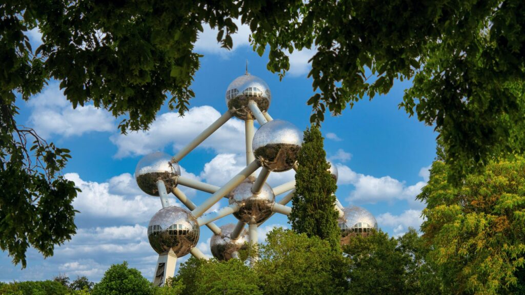 The Atomium, a large stainless steel structure of nine interconnected spheres representing an iron crystal lattice, rises above a canopy of green trees against a blue sky with white clouds. The reflective metallic spheres are connected by cylindrical tubes, with lush tree branches framing the structure on all sides.