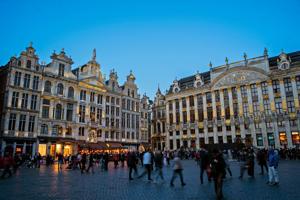 Brussels' Grand-Place at dusk, with illuminated Baroque and Gothic guild houses lining the cobblestone square as crowds of visitors move through the blue-hour light. Gold-ornamented stone façades with stepped and curved gables rise against a deep blue sky, with ground-floor shops and cafés glowing warmly in the foreground.
