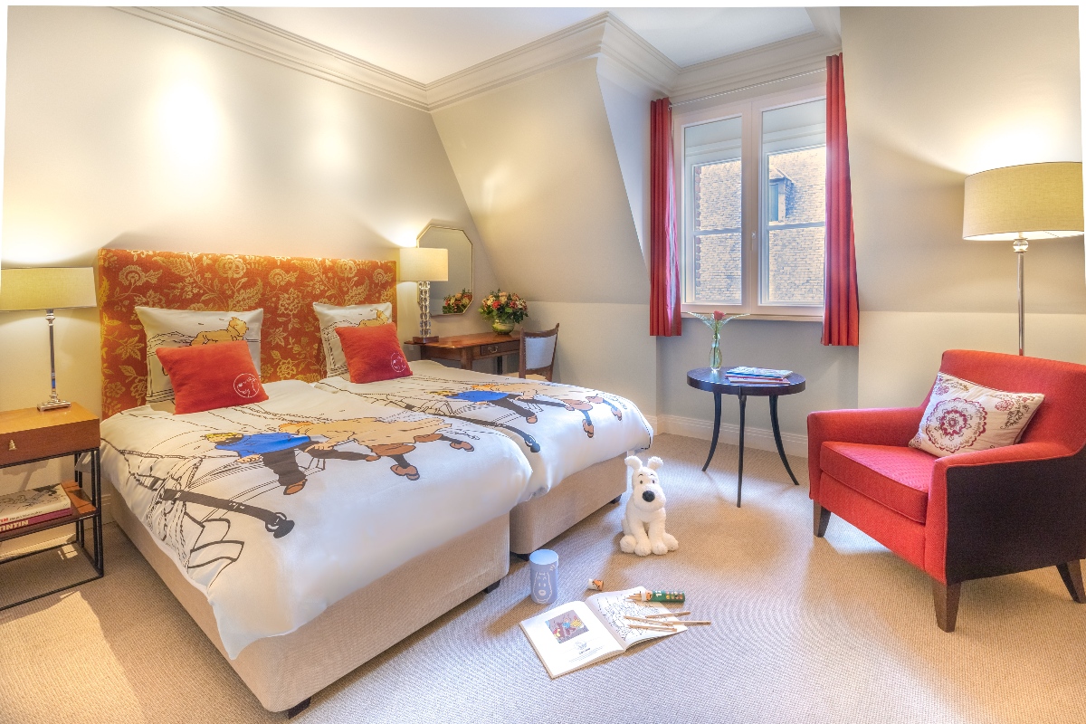 A hotel room with a double bed featuring a white duvet printed with Tintin comic strip characters, an orange floral upholstered headboard, and red accent pillows, with a white plush dog toy, open coloring book, and colored pencils arranged on the floor. The room includes red curtains framing a window, a red armchair, warm lamp lighting, and a small round side table with a flower arrangement.