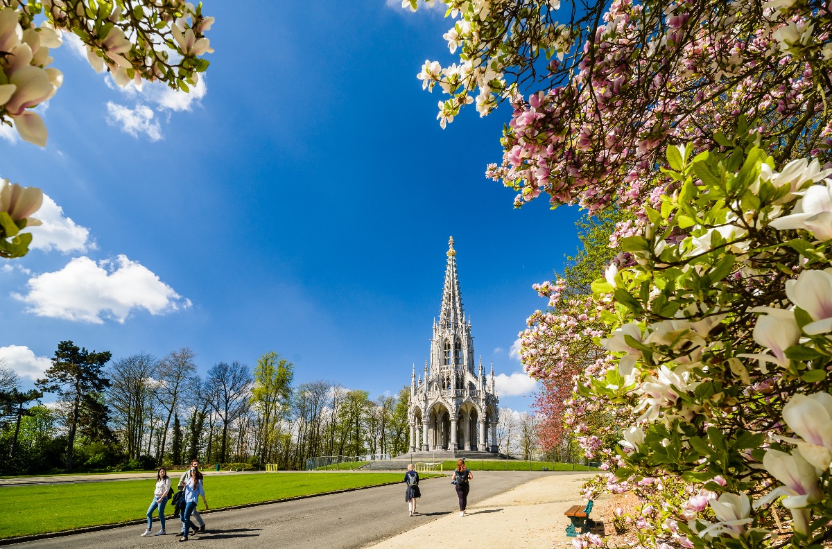A tall white Gothic spire monument rises at the end of a broad park pathway, flanked by green lawns and bare-branched trees, as several visitors stroll in the foreground. Pink and white magnolia blossoms frame the right side and upper corners of the image against a bright blue sky with scattered clouds.