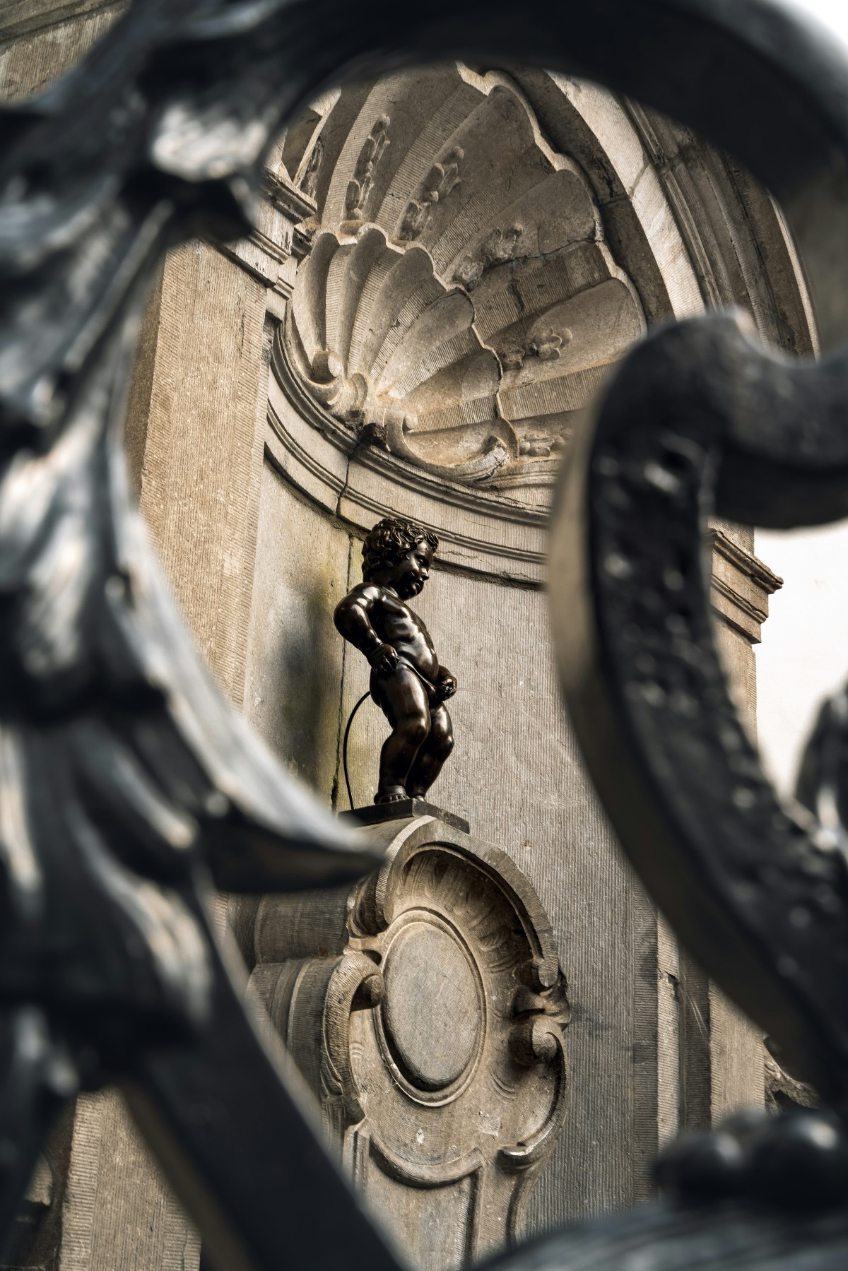 The Manneken Pis, a small dark bronze sculpture of a urinating boy, stands on a carved stone pedestal set into an ornate limestone niche with a shell-shaped relief above. The statue is photographed through the blurred curves of a decorative iron gate in the foreground, with the stone fountain structure's medallion and architectural detail visible below.