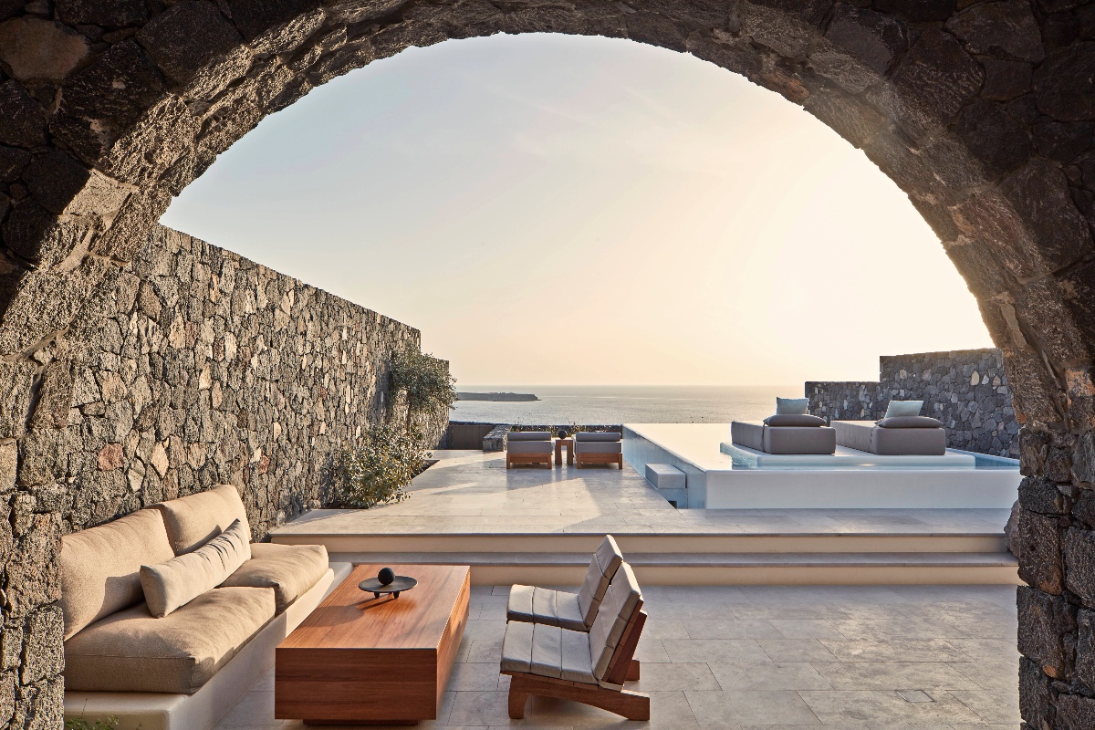 Outdoor terrace of a two-bedroom pool villa at Canaves Epitome, Santorini, viewed through a large volcanic stone arch, with a built-in linen sofa and teak coffee table in the foreground and a stepped infinity pool terrace with teak lounge chairs beyond. Dry-stacked volcanic stone walls frame the space on either side, with an unobstructed view of the Aegean Sea and a small island on the horizon under a pale sunset sky.