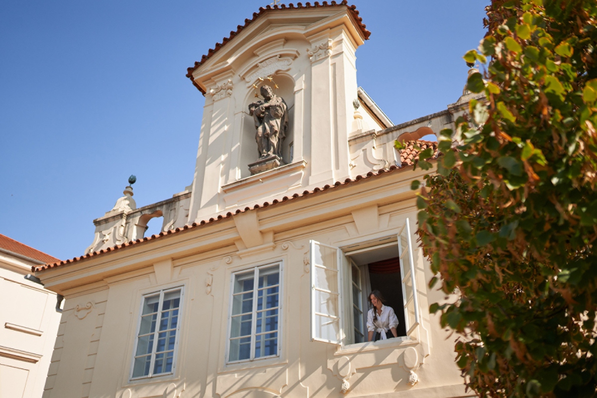 Exterior of the 16th-century Baroque Riverside Villa at Four Seasons Hotel Prague, photographed from below, showing the white stucco facade with terracotta roof tiles, ornate Baroque pediment, and a stone religious statue set into a niche above the roofline. A woman leans out of an open casement window on the upper floor, with a green-leafed tree visible in the right foreground against a clear blue sky.