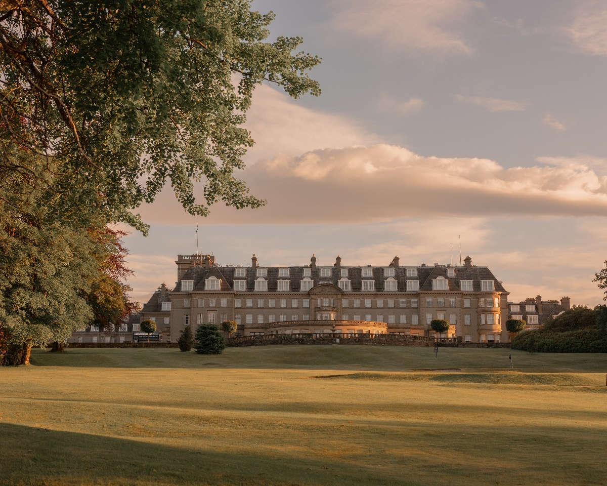 The Gleneagles Hotel in Perthshire, Scotland, photographed from the golf course at golden hour, showing the full facade of the five-story Edwardian sandstone building with its distinctive mansard roof, dormer windows, and central portico entrance. The hotel is framed by mature deciduous trees in early autumn foliage on the left, with manicured fairway in the foreground and a warm-toned cloudy sky above.