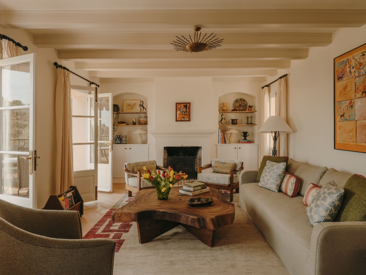 Living room of Villa Robert Graves at La Residencia, A Belmond Hotel, Mallorca, featuring a live-edge walnut coffee table, a linen sofa with mixed textile pillows, and two wood-framed armchairs flanking a white plaster fireplace, with built-in arched niches displaying ceramics and local artwork on either side. Exposed beam ceilings, terracotta tile floors, a sunburst ceiling fixture, and a large figurative painting on the right wall reflect the Studio Ashby renovation's integration of Mallorcan craft and the hotel's art collection, with French doors open to a sunlit terrace beyond.