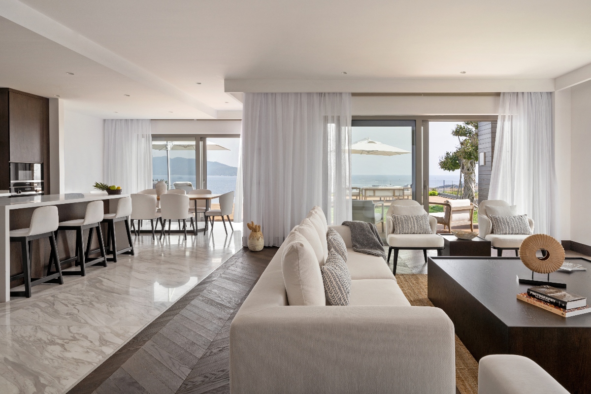 Open-plan living and dining area of Villa Poseidon at METT Bodrum, featuring a cream linen sectional sofa, dark wood coffee table, and marble-effect tile floors, with a kitchen island and bar seating visible on the left and a dining table with upholstered chairs beyond. Floor-to-ceiling sliding glass doors on two sides open to furnished terraces with market umbrellas, with panoramic views of the Aegean Sea and the hills of the Bodrum Peninsula in the background.