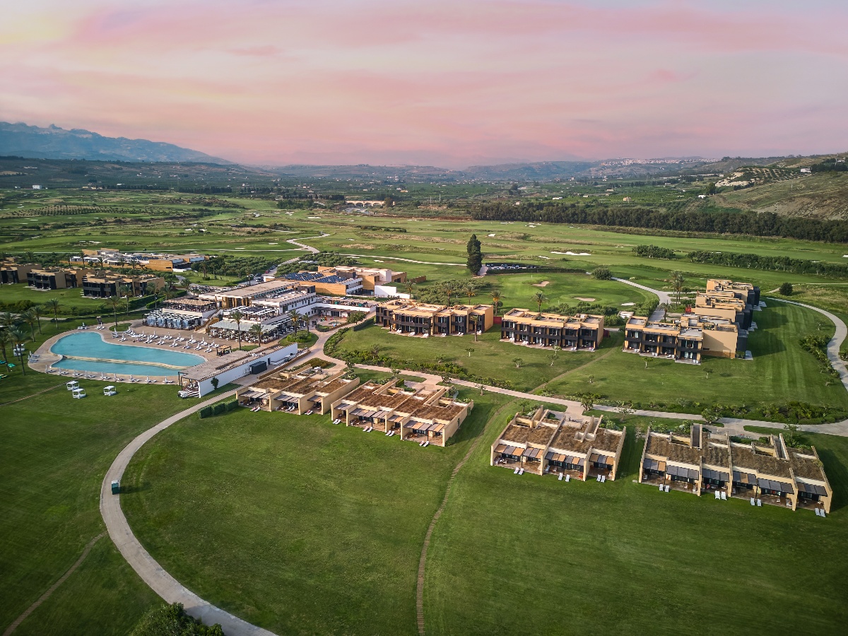 Aerial dusk view of Verdura Resort on Sicily's southwestern coast, showing the main hotel complex, a large freeform pool with lounge chairs, and rows of low-rise terracotta-toned villas with private terraces distributed across manicured grounds. A championship golf course extends across the surrounding landscape, with rolling Sicilian hills, olive groves, and a pink-tinged sky visible in the background.