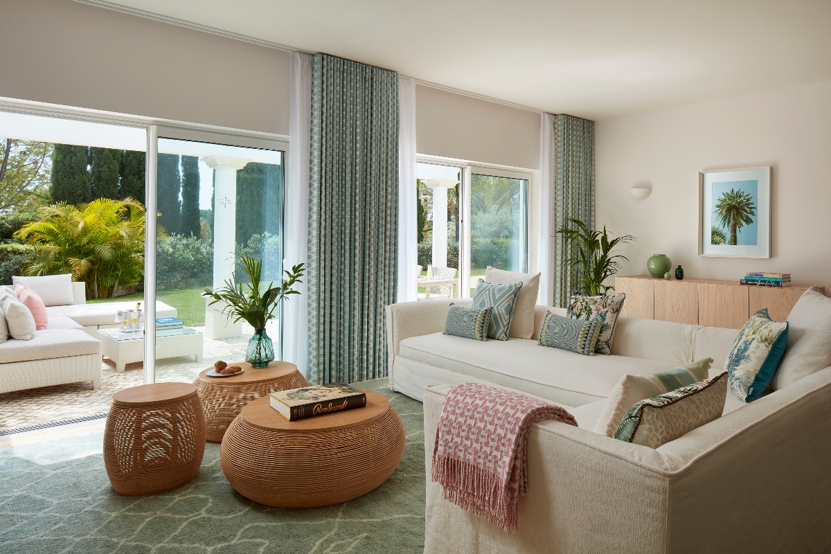 Living room of a Vila Vita Parc resort villa, featuring two white linen sofas with teal and pink accent pillows, a pink throw, and a pair of round rattan ottoman coffee tables on a sage green patterned rug. Floor-to-ceiling sliding glass doors open directly onto a furnished terrace and lush garden with palm trees and tropical plantings, with a framed coastal print and oak sideboard visible on the interior wall.