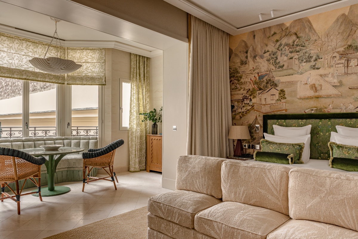 Junior suite or apartment interior at Villa Saint James, Paris, featuring a king bed with a deep green velvet headboard and botanical-print pillows set against a large-scale chinoiserie landscape mural, with a cream textured sofa in the foreground. An open-plan dining area with a green pedestal table, rattan and woven chairs, and botanical roman shades adjoins the sleeping space, with French doors opening to a wrought-iron balcony.
