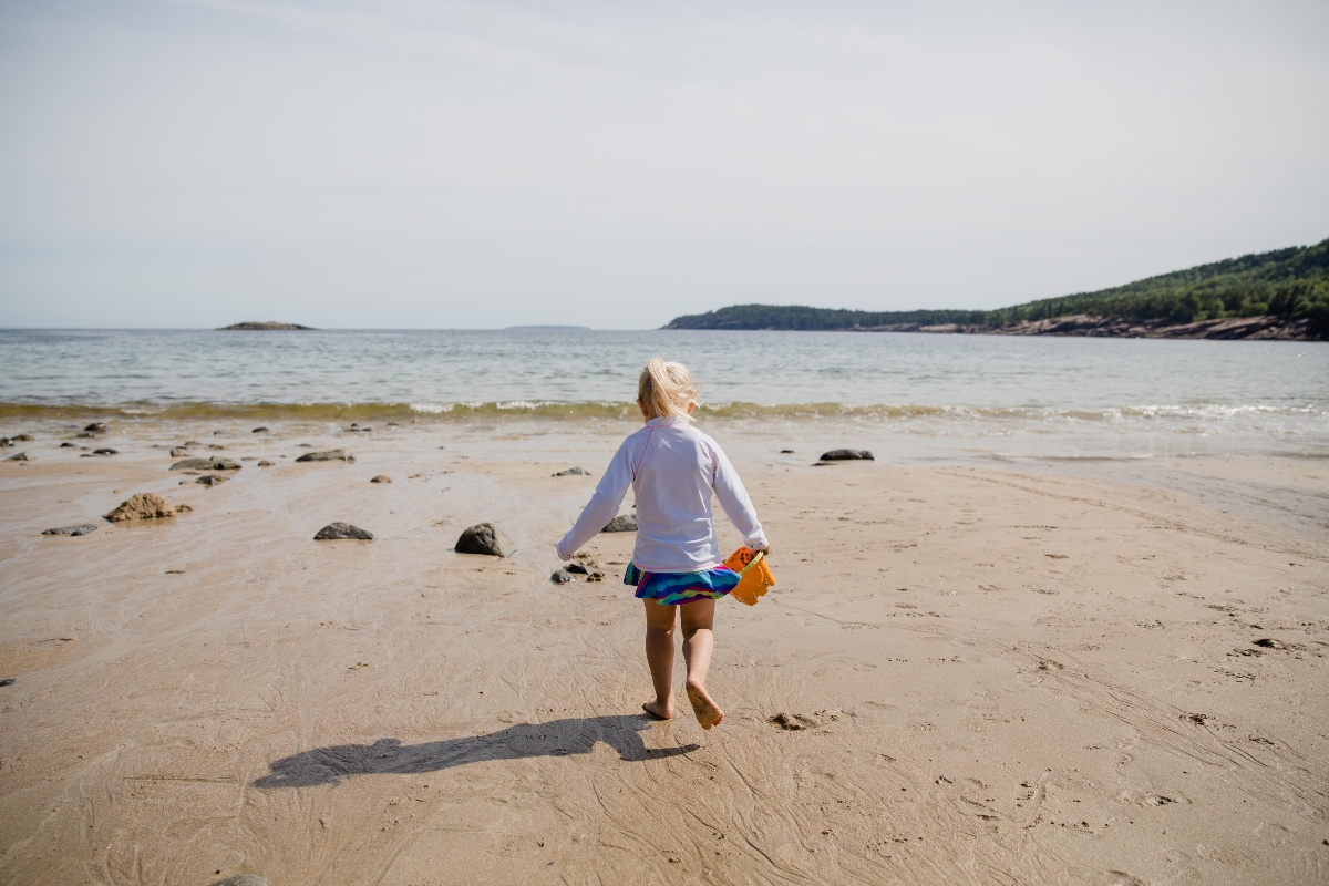 A young child with blonde hair runs barefoot toward the water on a sandy beach, wearing a white long-sleeve rashguard, a colorful skirt, and carrying an orange item in one hand. Rocky outcroppings, a forested headland, and a small island dot the calm shoreline in the background under an overcast sky.
