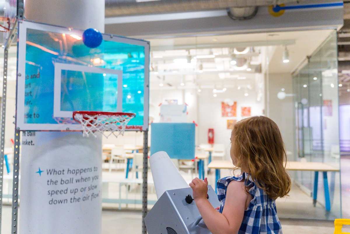 A young child with curly brown hair stands at an air-powered launcher at a children's museum exhibit, directing a blue ball toward a small basketball hoop mounted on a backboard. A sign on the nearby column reads "What happens to the ball when you speed up or slow down the air flow?" suggesting an interactive science activity.