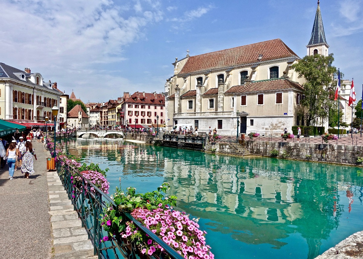 A green iron railing lined with pink flowering plants borders a canal of vivid turquoise water, with pedestrians walking along the quay on the left and a large cream-colored church with a terracotta roof and pointed steeple reflected in the water on the right. Colorful multi-story buildings, a stone bridge, and French and Swiss flags are visible in the background under a partly cloudy blue sky.