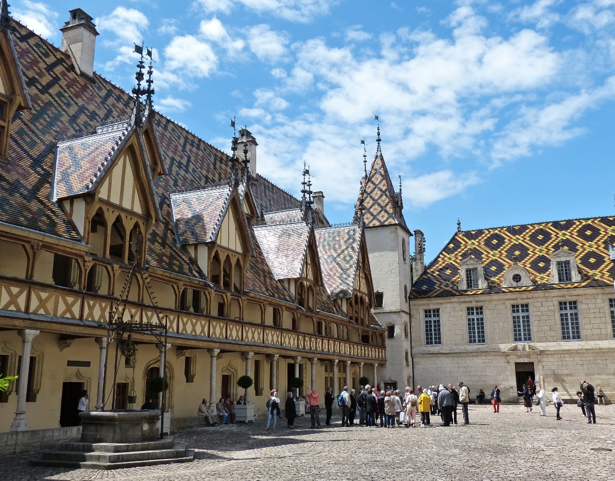 The cobblestone inner courtyard of the Hospices de Beaune is framed by Gothic timber-framed buildings with steep, multicolored glazed tile roofs patterned in geometric designs of black, gold, and rust. A group of visitors gathers near the entrance of the adjoining stone building on the right, while an ornate iron well stands along the arcade on the left.
