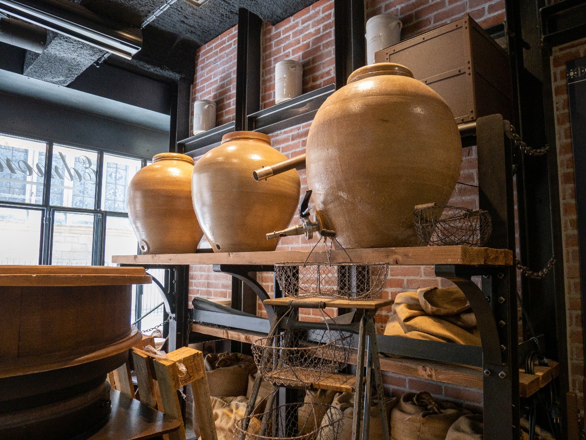 Three large round stoneware crocks fitted with brass spigots and spouts sit on a wooden-planked metal shelving unit against an exposed brick wall, with wire mesh baskets and burlap sacks visible on the lower shelves. A large window with frosted lettering admits natural light from the left, illuminating the warm amber tones of the ceramic vessels in what appears to be a production or display space.