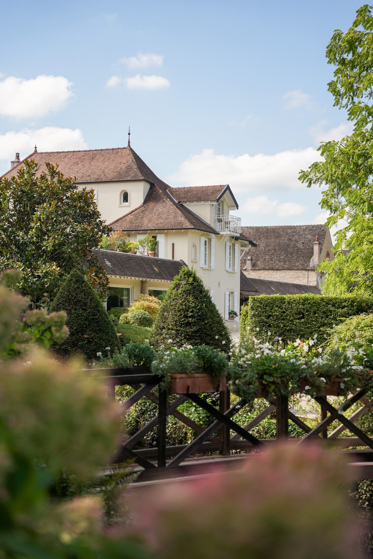 A cream-colored French manor house with steeply pitched terracotta tile roofs rises above manicured topiary and lush garden hedgerows, viewed through soft foreground blooms on a sunny day. Geometric cone-shaped shrubs, a wooden lattice fence with climbing flowers, and layered greenery frame the property's multi-building facade beneath a partly cloudy blue sky.