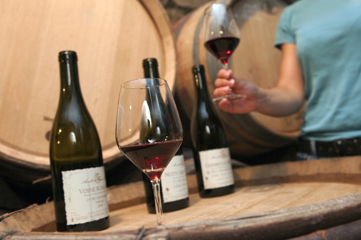 Three dark green wine bottles with white labels, including a visible "Vosne-Romanée" and "Nuits-Saint-Georges," are arranged on a large oak barrel alongside a partially filled red wine glass in a barrel-lined cellar. In the background, a person in a light blue shirt holds a second glass of red wine up for examination.
