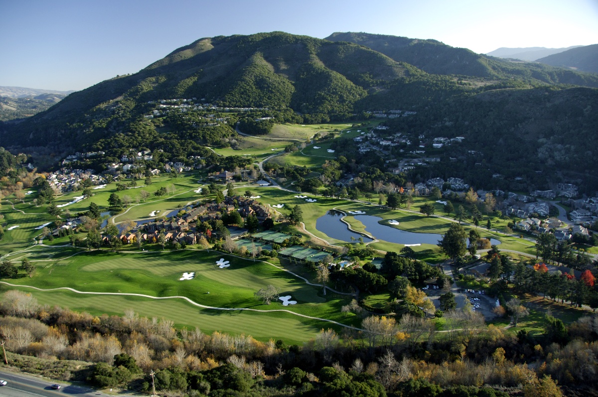 An aerial view of a resort property nestled in a broad valley, featuring a golf course with sand bunkers, a pond, tennis courts, and lodge-style buildings surrounded by mature trees showing autumn foliage. Rolling green hills rise steeply on multiple sides, with a residential community and winding roads visible along the valley slopes beneath a clear blue sky.