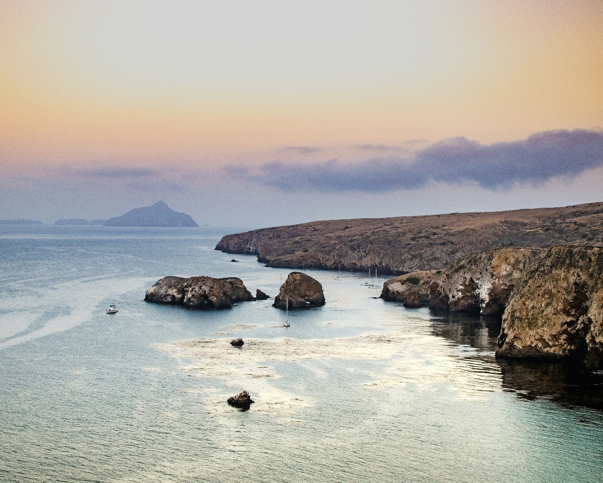 An elevated view of a rocky island coastline at dusk, with scattered rock formations and several sailboats anchored in calm, shallow water below rugged tan-and-gray cliffs. A soft gradient sky in shades of peach, lavender, and gray stretches above low coastal fog, with the silhouette of a distant island visible on the horizon.