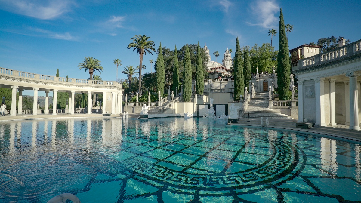The Neptune Pool at Hearst Castle features a large outdoor swimming pool with a detailed mosaic tile pattern visible through the clear blue water, flanked by white marble colonnades, classical statuary, and grand staircases. Tall Italian cypress trees and palm trees frame the grounds, with the castle's ornate Spanish-Mediterranean towers visible in the background beneath a clear blue sky.