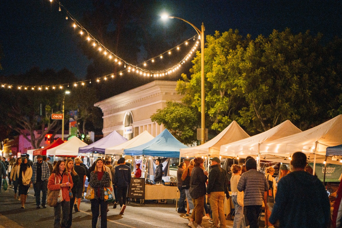 A busy outdoor farmers market fills a closed downtown street at night, with vendors under white and colorful canopy tents lining both sides as crowds of people browse in casual clothing. String lights arc overhead against a dark sky, with a classical-style building facade and large trees visible in the background.