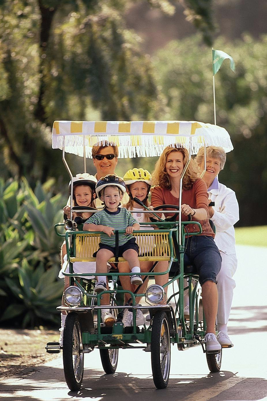 A multi-generational group of three adults and three helmet-wearing children ride a pedal-powered surrey bike along a tree-lined paved path on a sunny day. The four-wheeled surrey features a yellow-and-white striped fringed canopy and a small green flag, with the passengers smiling toward the camera.