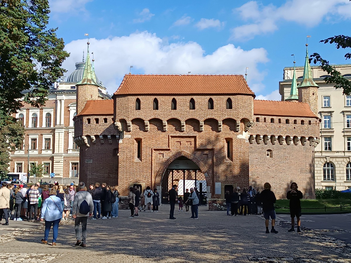 The Kraków Barbican, a late 15th-century red brick fortified gatehouse with a central arched entrance, crenellated walls, and orange-tiled roofs, stands against a blue sky with scattered clouds. A cobblestone plaza in front of the structure is filled with visitors, with historic multi-story buildings visible on either side.