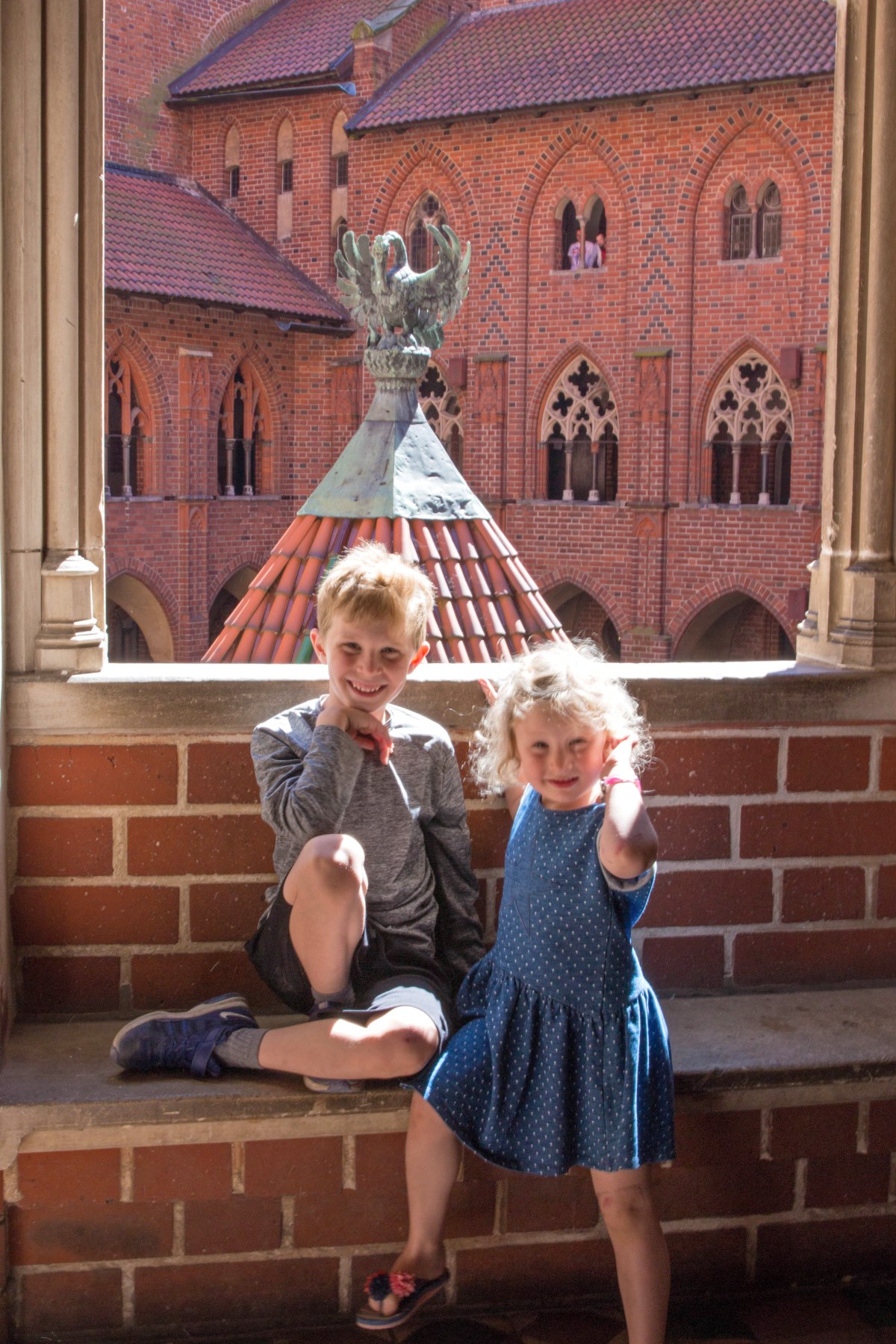 Two children sit in a stone window opening at Malbork Castle, with the red brick Gothic architecture of the castle's inner courtyard visible behind them. A bronze eagle sculpture crowns a conical roof in the background, and arched Gothic windows line the castle wall beyond.
