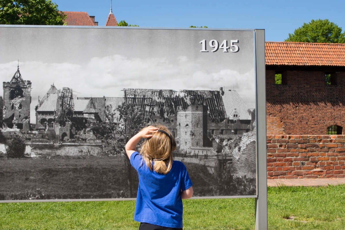 A young girl with her back to the camera stands on a grass lawn examining a large outdoor display panel showing a black-and-white photograph of a heavily damaged castle labeled "1945." The restored red brick walls of Malbork Castle are visible to the right, providing a present-day contrast to the wartime destruction depicted in the historical image.