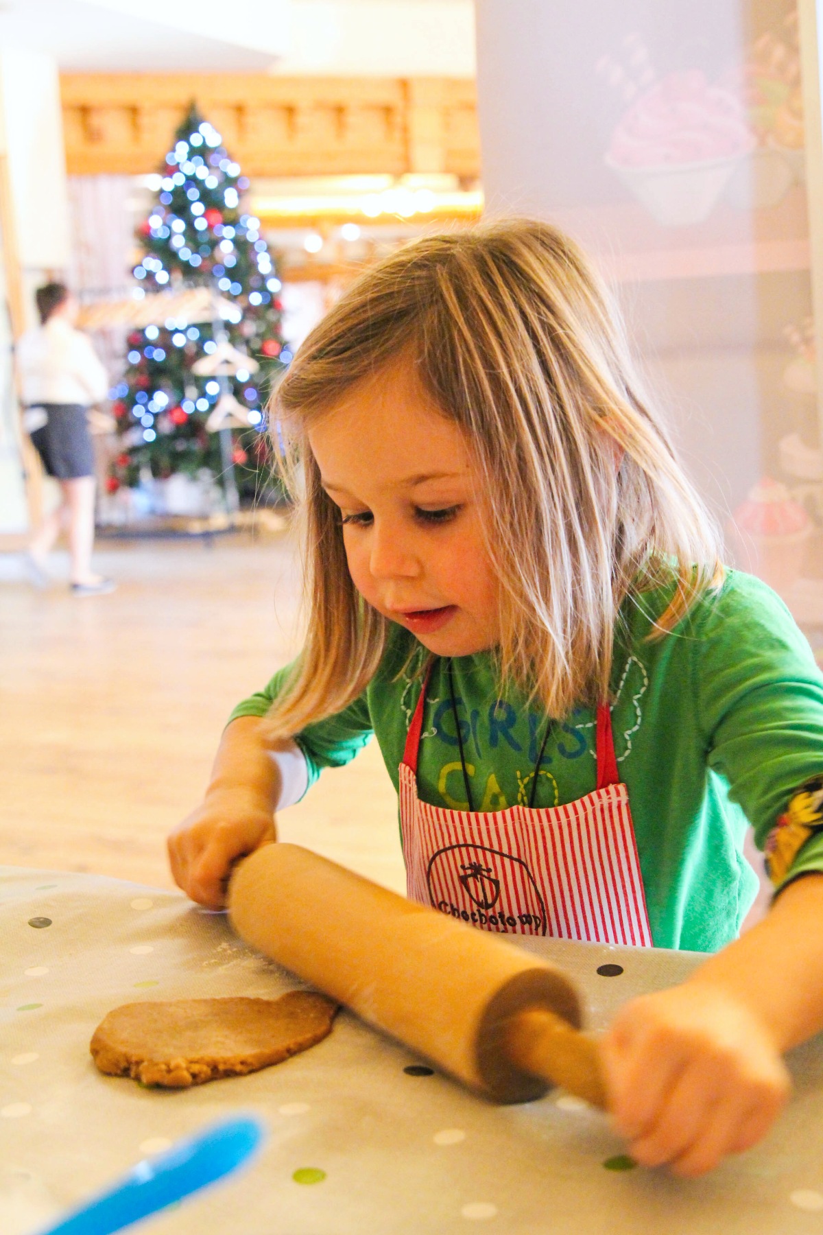 A young girl wearing a red-and-white striped apron over a green shirt rolls out dough with a wooden rolling pin on a flour-dusted surface. A decorated Christmas tree glows in the background of the bright, open space behind her.