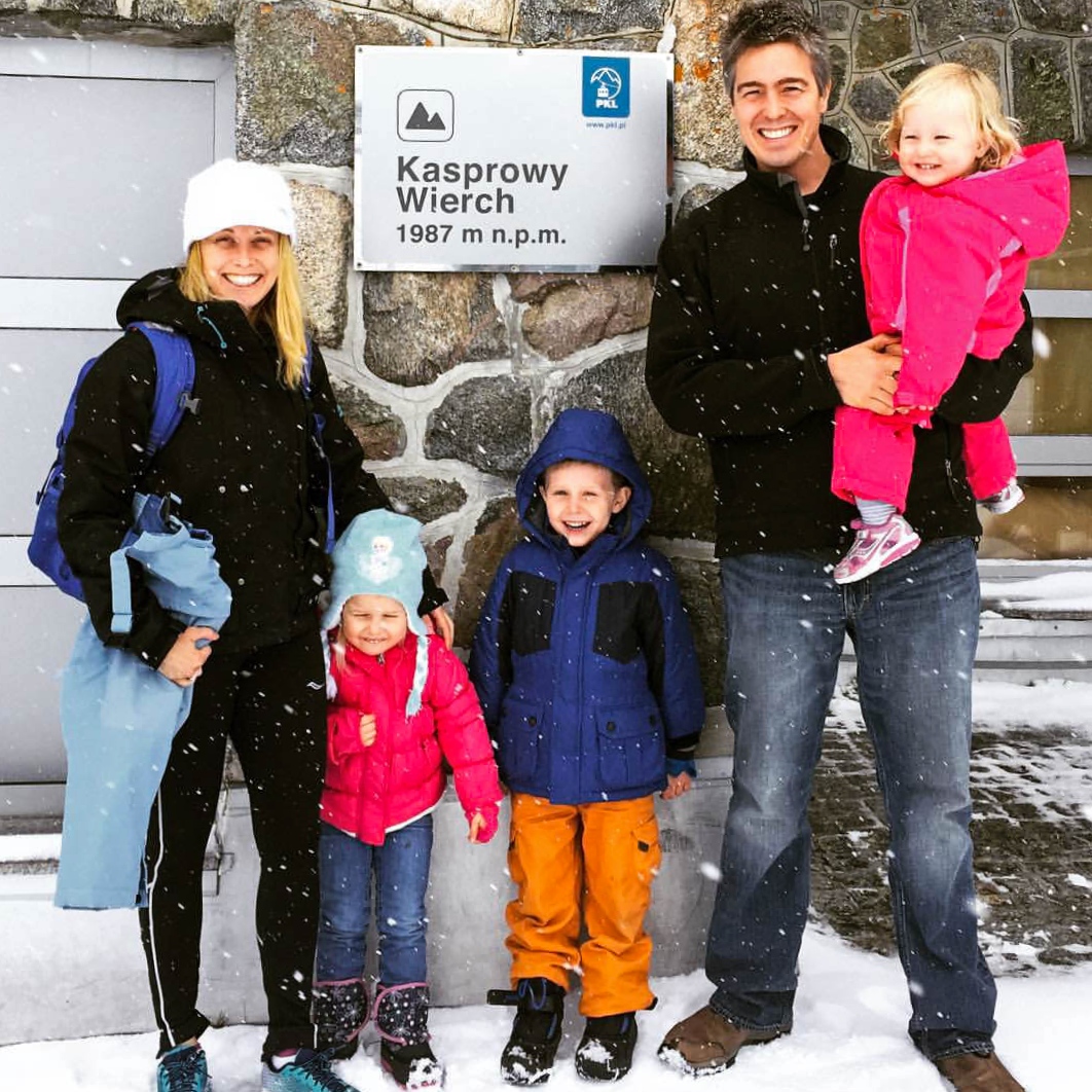 A family of five poses in falling snow beside a stone wall-mounted sign reading "Kasprowy Wierch 1987 m n.p.m." in the Tatra Mountains of Poland. Two adults and three young children, dressed in winter jackets and hats, smile at the camera, with the PKL cable car logo visible on the sign above them.