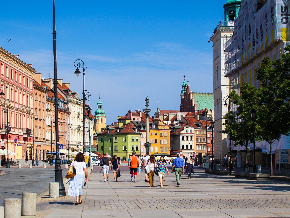 Pedestrians walk along a broad, sun-lit promenade toward Warsaw's Castle Square, where colorful historic buildings in shades of yellow, green, and pink line the plaza beneath a clear blue sky. A tall column monument — Sigismund's Column — rises at the center of the square, flanked by church spires and green copper-topped towers in the background.