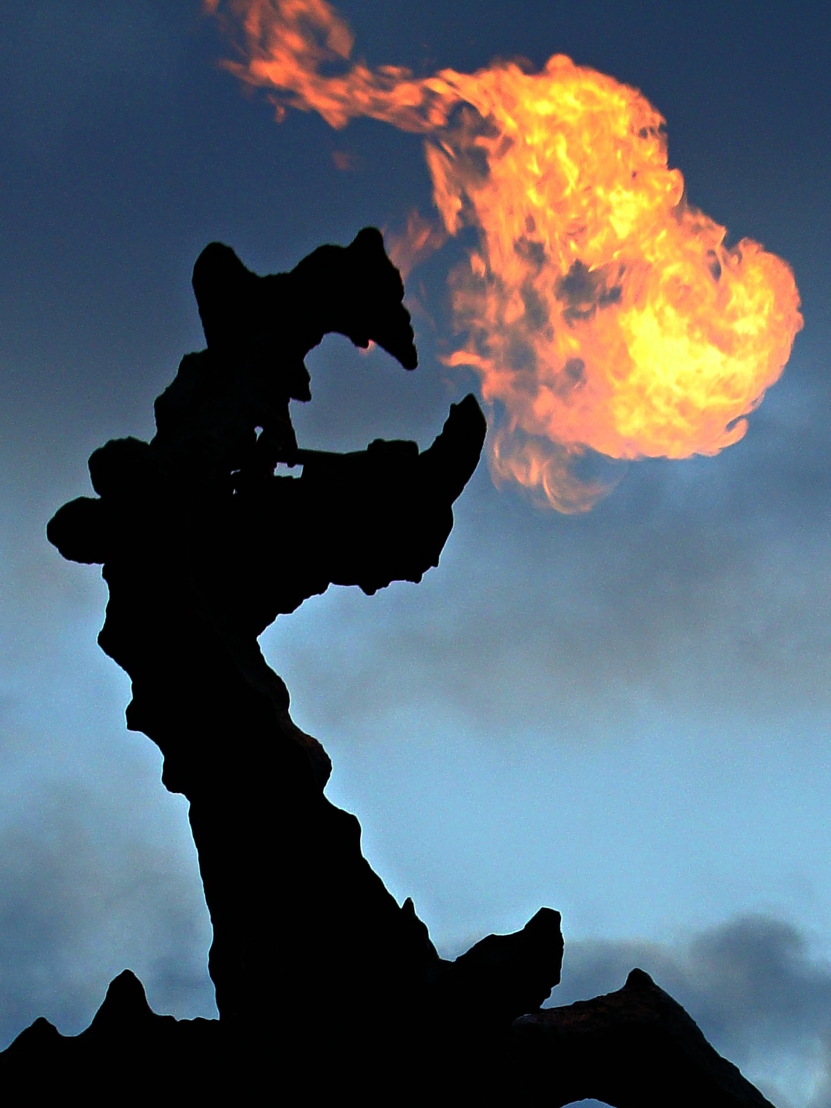 The bronze Wawel Dragon statue is silhouetted in near-total darkness against a dusky blue sky, with a large burst of real flame erupting from its open mouth. The sculpture, which stands at the base of Wawel Hill in Kraków, periodically breathes fire powered by natural gas.