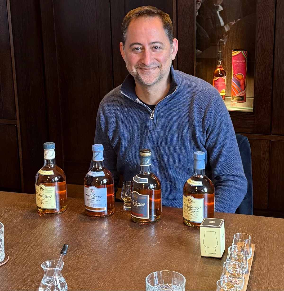 A smiling man in a blue quarter-zip sweater sits at a wooden table during a Dalwhinnie whisky tasting, with four bottles of single malt Scotch, a tasting glass, and a small water jug arranged in front of him. Behind him, a display case holds additional bottles against dark wood paneling, and several nosing glasses sit on a wooden flight tray in the foreground.