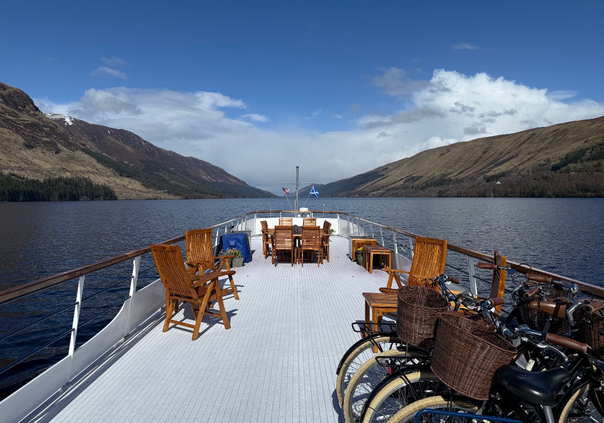 The sun deck of a hotel barge holds wooden folding chairs around a dining table and a row of bicycles with wicker baskets lined up along the right railing, with an American flag and the Scottish saltire flying from a mast at the bow. Beyond the deck, a loch stretches between steep green and brown hillsides under a blue sky scattered with clouds.