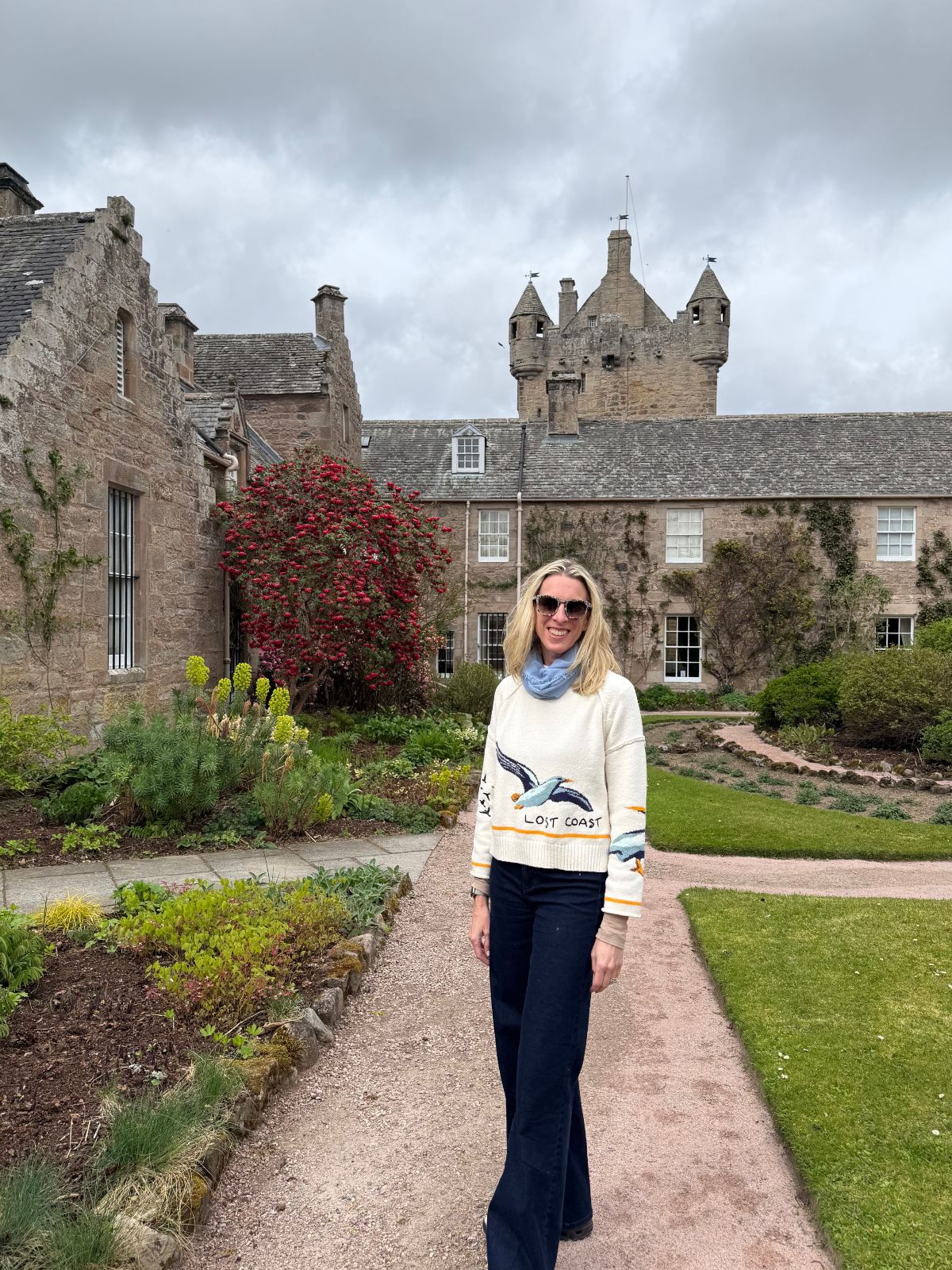 A blonde woman in sunglasses, a light blue scarf, a cream "Lost Coast" sweater with a bird design, and dark flared jeans stands on a gravel path in the garden of a stone castle estate. Behind her, a turreted castle and adjoining stone buildings rise above planted flower beds, a manicured lawn, and a flowering red shrub under an overcast sky.