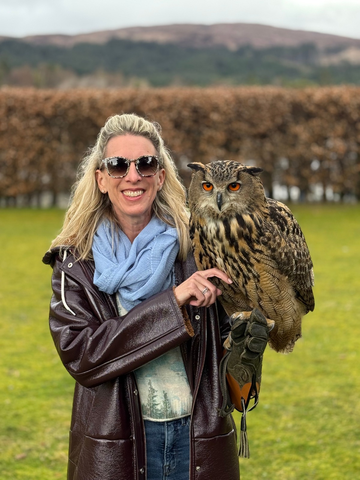 A smiling woman in sunglasses, a light blue scarf, and a brown leather coat holds a large eagle owl with orange eyes and mottled brown plumage perched on her gloved hand. Behind her, a manicured lawn, a tall trimmed hedge, and a brown hillside extend under an overcast sky.