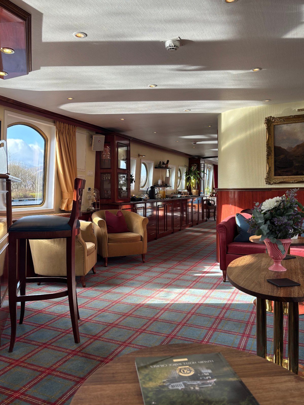 The lounge of a luxury hotel barge features a blue and red tartan carpet, mahogany cabinetry with a glass-fronted cabinet of stemware, and tan and burgundy upholstered armchairs arranged near arched windows. A round wooden table in the foreground holds a pink vase of white and purple flowers, with a framed landscape painting on the wall and a "Luxury Hotel Barge Cruises" brochure resting on a table at the bottom of the frame.