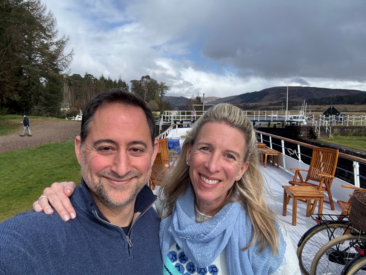 A man in a blue quarter-zip sweater and a woman in a light blue scarf and patterned sweater smile for a selfie on the sun deck of a hotel barge, with wooden folding chairs and bicycles visible behind them. Beyond the deck, a canal lock, moored boats, evergreen trees, and brown hills stretch under a cloudy sky.