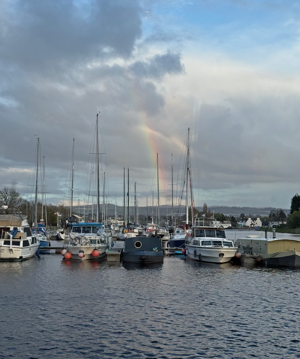 A rainbow descends through gray and blue clouds over a marina where sailboats, motorboats, and a dark canal barge are moored in calm water. Low wooded hills and a cluster of white buildings line the far shore beneath an overcast sky.