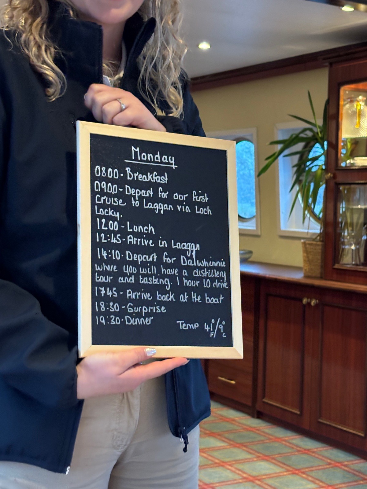 A crew member in a black jacket holds a wood-framed chalkboard displaying the Monday schedule for a barge cruise, with handwritten times listing breakfast, a cruise to Laggan via Loch Lochy, lunch, a Dalwhinnie distillery tour and tasting, dinner, and a noted temperature of 41°F/9°C. Behind her, the barge interior shows a tartan carpet, mahogany cabinetry with a glass-fronted cabinet, a potted plant, and arched windows.
