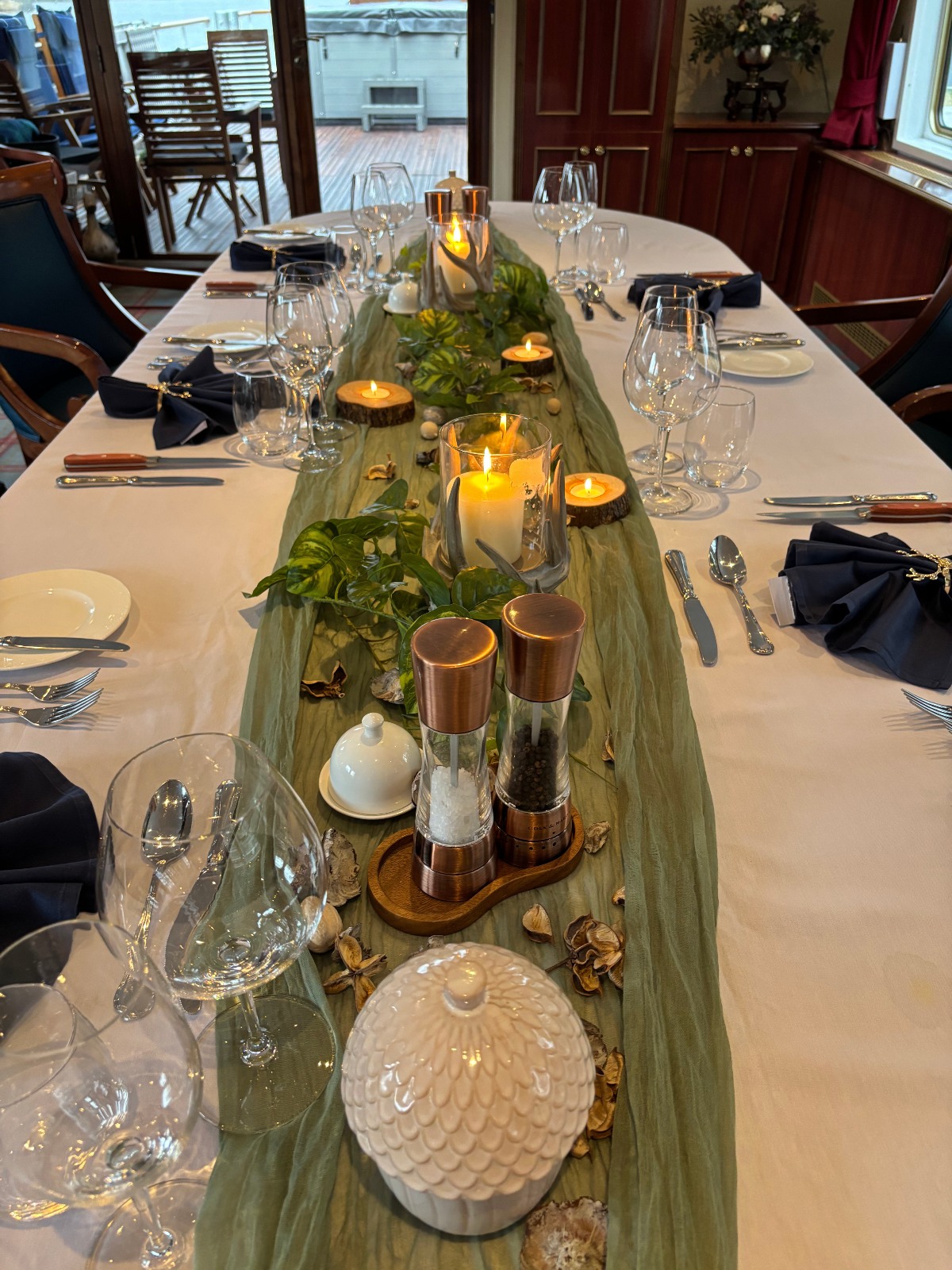 A long dining table set with white linens, navy napkins, multiple wine glasses, and white plates runs through the dining saloon of a hotel barge, with a sage green runner trailing candles, greenery, dried botanicals, and wood-slice coasters down the center. Copper salt and pepper mills, a white artichoke-shaped tureen, and a butter dish anchor the foreground, while an open doorway at the rear reveals the sun deck and its wooden furniture.