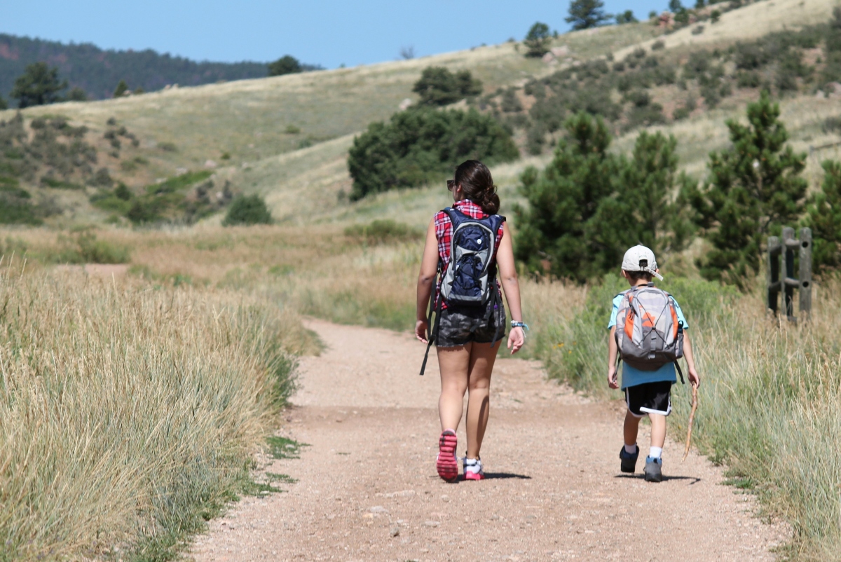 A woman and a young boy walk along a gravel trail through a grassy, hilly landscape dotted with pine trees, both carrying backpacks and facing away from the camera. The boy wears a white baseball cap, blue shirt, and black shorts and holds a stick, while the woman wears a plaid top, camouflage shorts, and sunglasses.