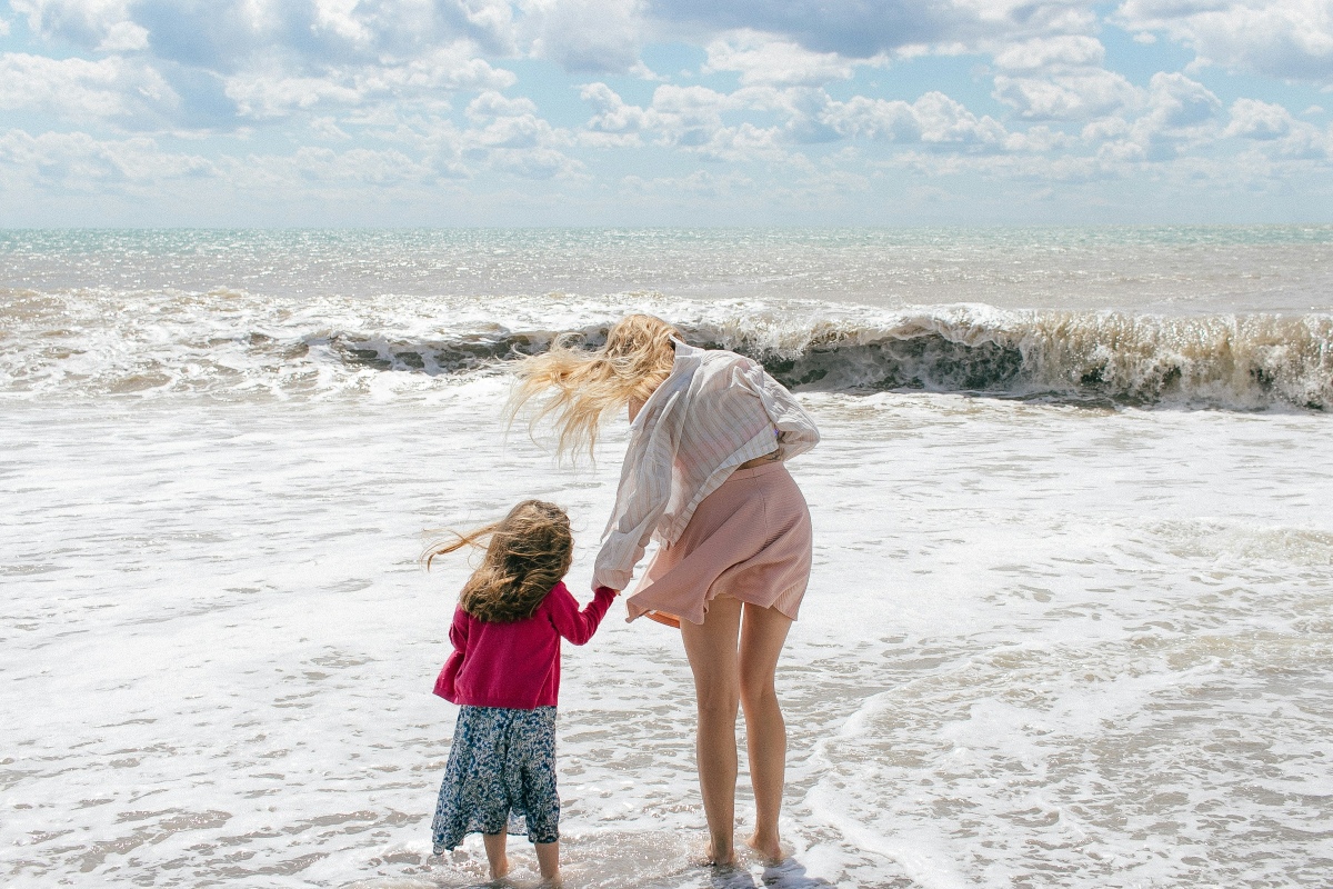 A woman and a young girl stand at the edge of the ocean holding hands, their hair blowing in the wind as waves roll in toward the shore. The woman wears a striped shirt over a pink skirt, while the girl wears a magenta cardigan over a blue floral skirt, both facing away from the camera toward the water.
